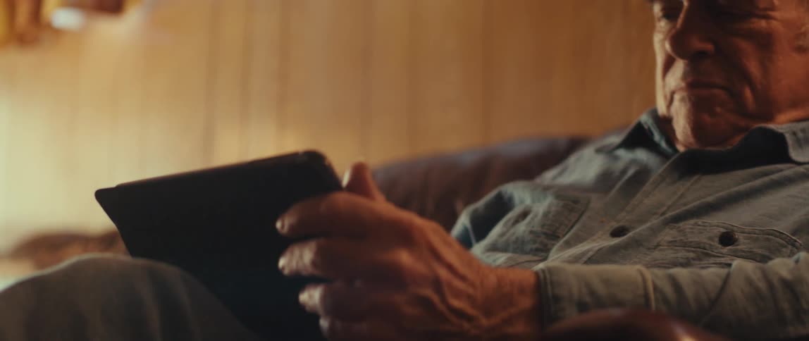 An elderly man in a cap and blue shirt sits on a leather couch using a tablet