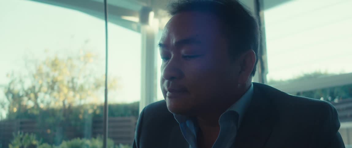 A man in a suit sits and looks thoughtfully out a large window before turning his attention to typing on a laptop