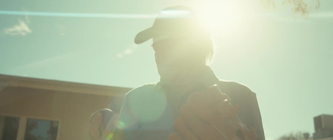 An elderly man wearing a baseball glove contemplates a baseball in his hand on a sunny day in the backyard