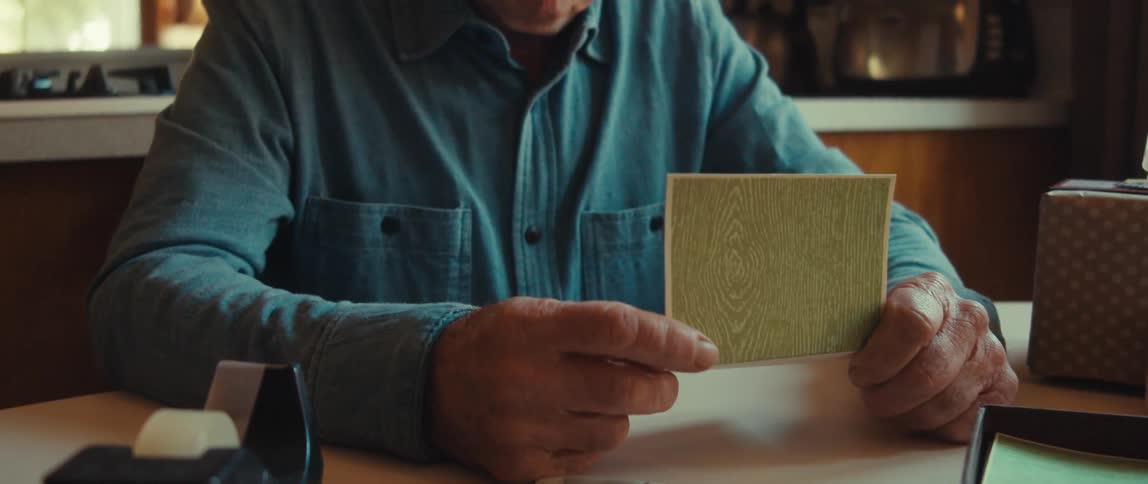 An elderly man is writing a note or a letter on a green card at a table