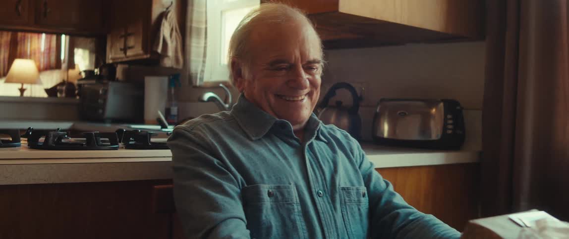 A happy elderly man holds a wrapped present while sitting in the kitchen at home