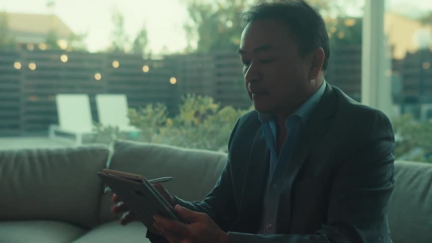 A man in a business suit sits on a couch at home using a tablet on a sunny morning
