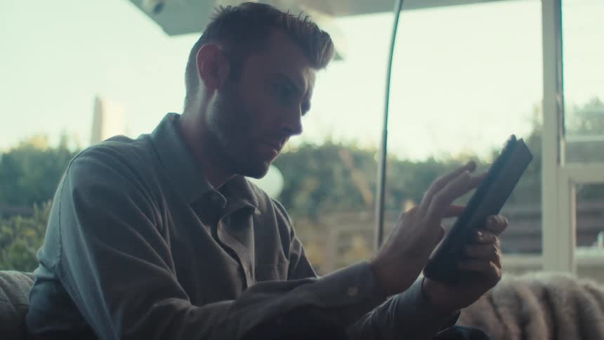 A young man sitting on a couch at home tapping on a tablet