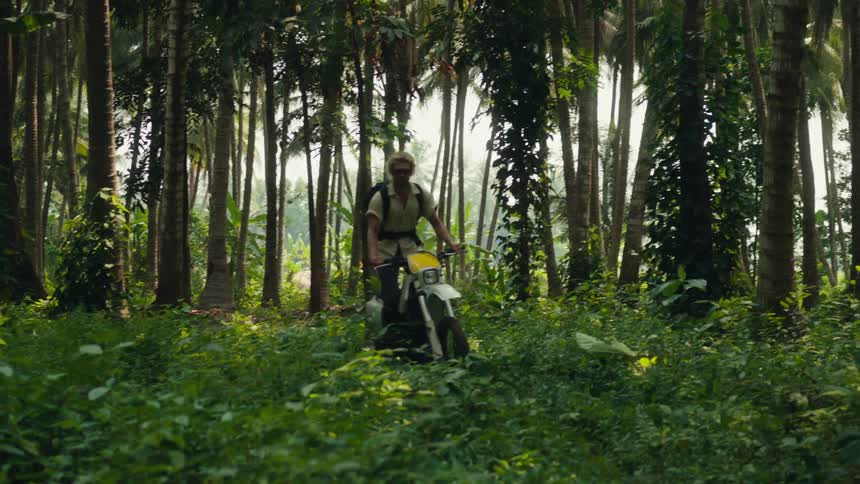 A man wearing a black backpack rides a dirt bike through a dense lush forest