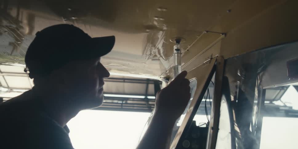 A person wearing a cap is inspecting fuel purity from the underside of an aircraft wing inside a hangar