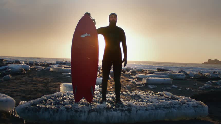A man stands on ice holding his surfboard