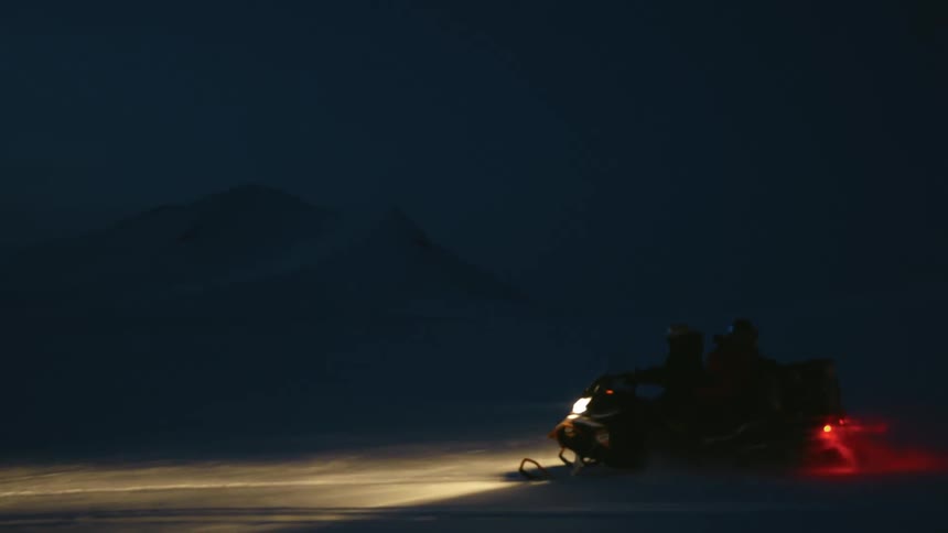 A snowmobile with two silhouetted figures on it is driving across a snowy dark landscape with its headlights and tail light illuminating the scene
