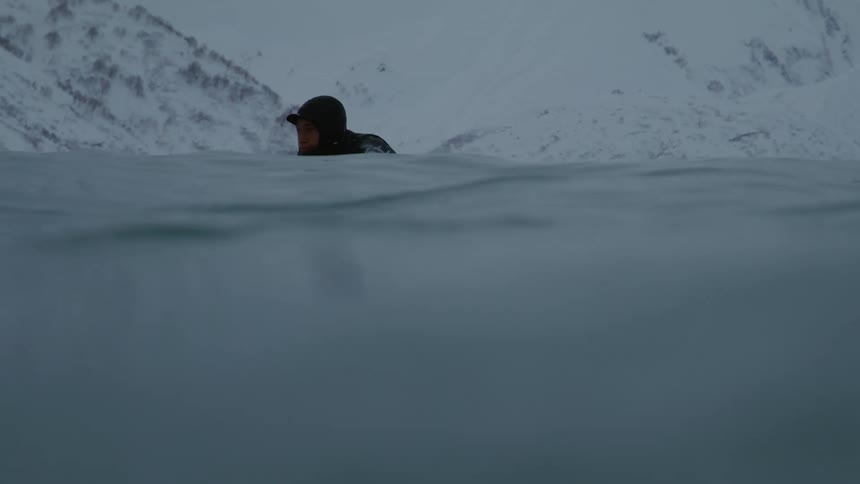 A person in a wetsuit paddles on a surfboard in cold water with snowcovered mountains in the background