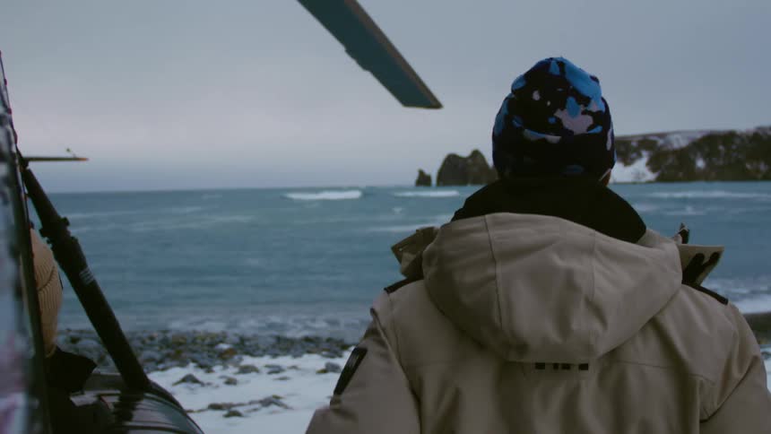 A person in a beige jacket and a blue patterned hat stands on a rocky snowdusted shore looking out at a cold choppy sea beside a helicopter