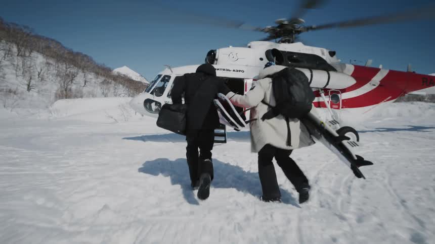 Two people carrying surfboards run to a helicopter while a person beckons them on a winter day