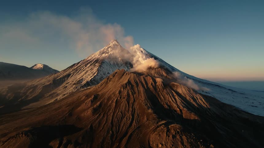 The peak of a huge mountain with smoke coming out of the top and behind there are other mountains with the top covered in snow