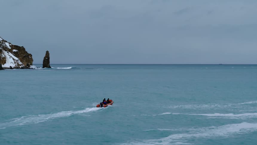 A small orange boat with three people is navigating through the calm turquoise waters near a rocky snowcovered coastline with a prominent sea stack