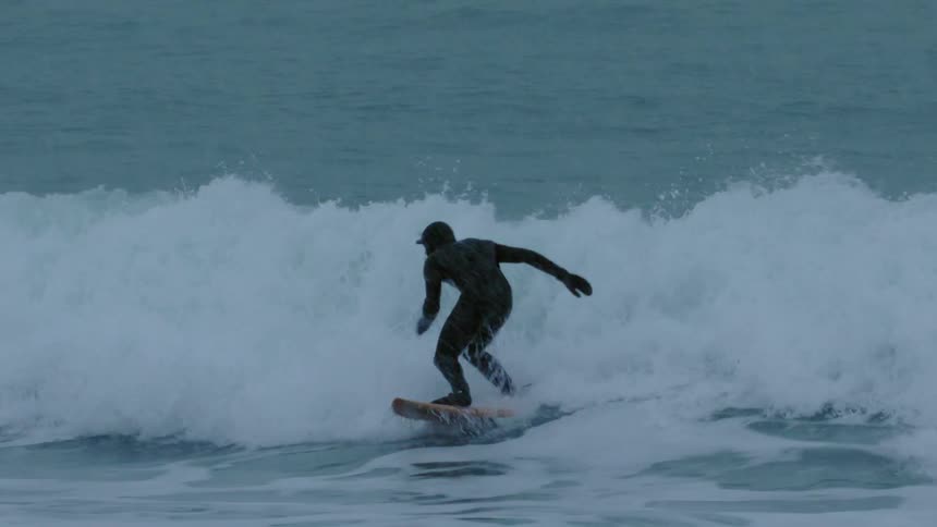 A surfer in a dark wetsuit rides a surfboard over waves near the coast