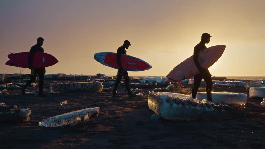 Three silhouetted surfers in wetsuits walk across a dark icy beach at sunset each carrying a surfboard