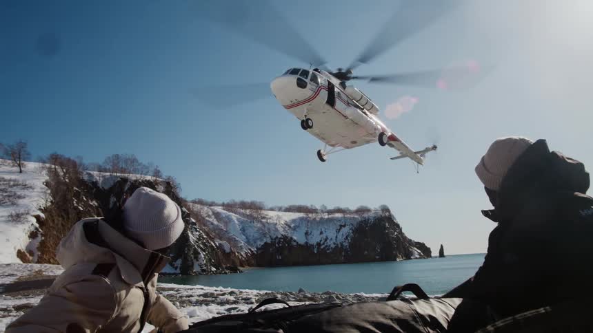 A white helicopter landing next to two people on the ground wearing warm clothing in a snowy environment
