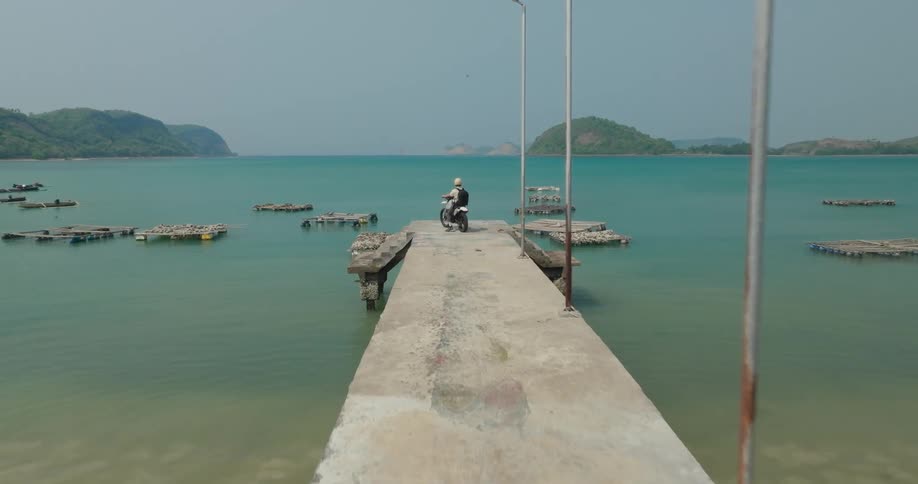 A person on a motorcycle rides to the end of a concrete pier extending into a turquoise sea