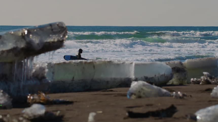 A surfer in a wetsuit carries a surfboard along a beach strewn with ice cubes