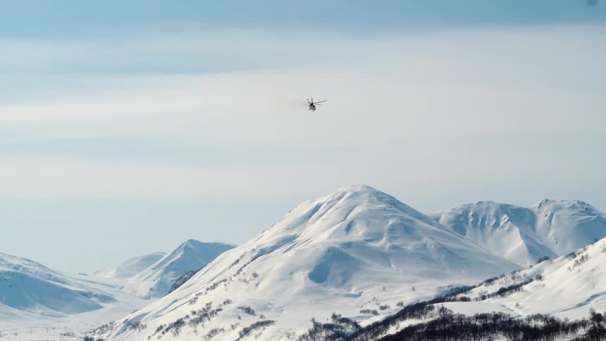 A helicopter flies over a snow capped mountain range with a clear blue sky above