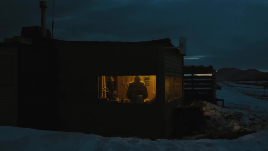 The image shows a small wooden cabin nestled in a snowy landscape with a lone figure silhouetted against the warm glow of the interior light