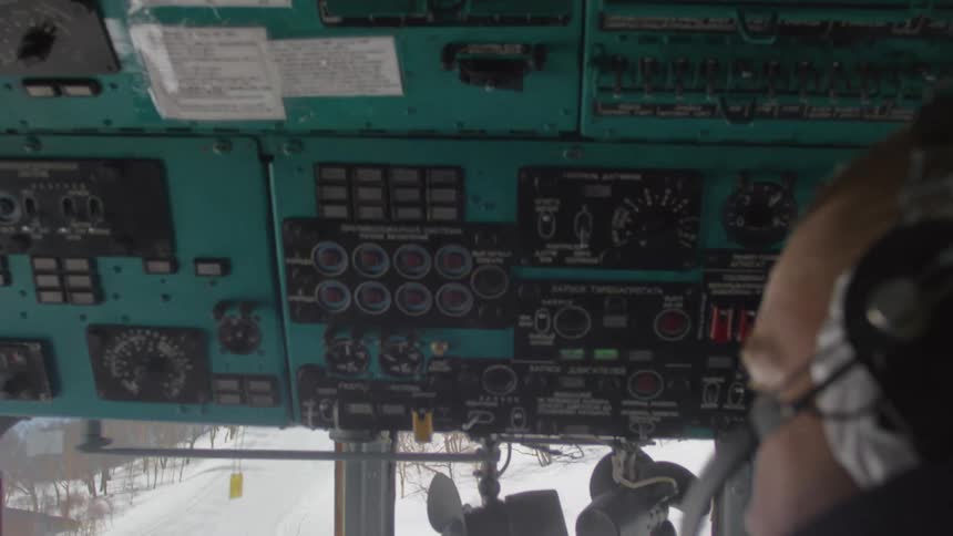 Two pilots on the interior of an aircraft cockpit with instruments and controls and a view through the windshield of a snowy landscape outside