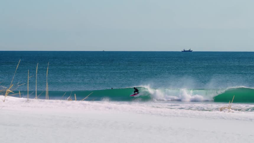 A surfer rides a perfect wave on a sunny day with a snowy beach in the foreground a calm ocean and a distant ship visible on the horizon