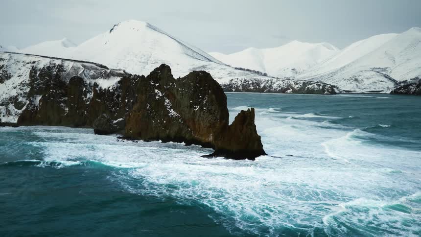 A rugged coastline with dramatic cliffs and snow capped mountains in the distance under a cloudy sky