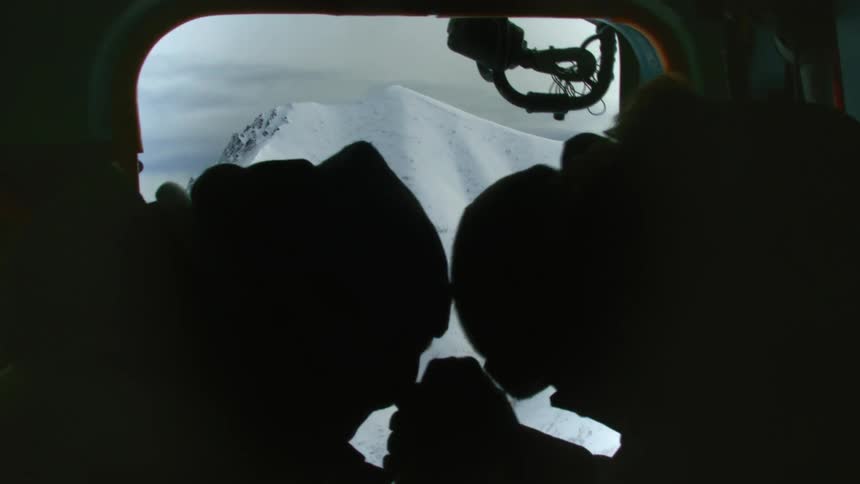 Two silhouetted figures inside an aircraft look out at a snow covered mountain through the window