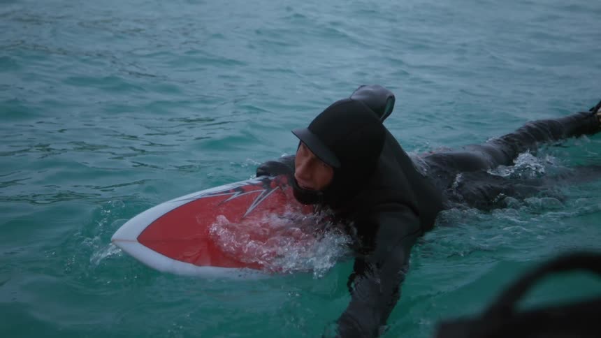 A person in a wetsuit paddles on a surfboard in cold turquoise water with snowcovered mountains in the background