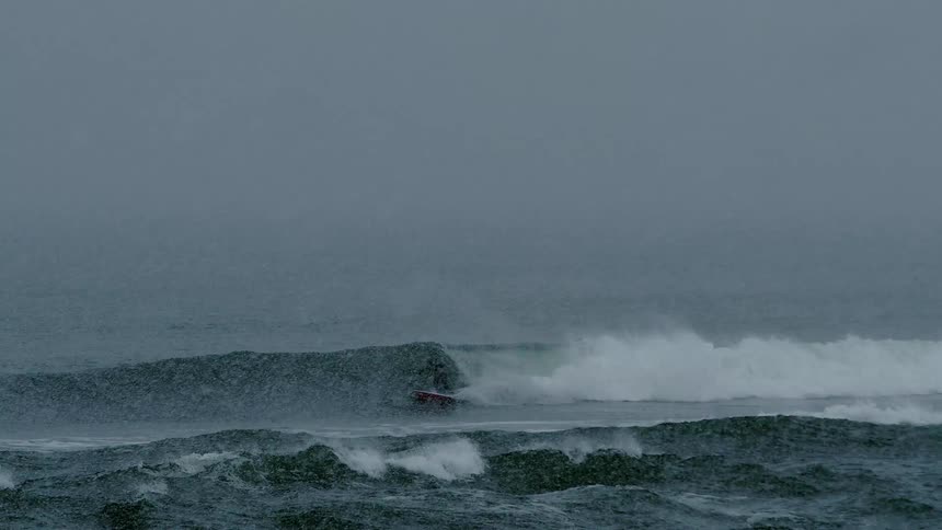 A surfer rides a wave near the coast during a light snowstorm