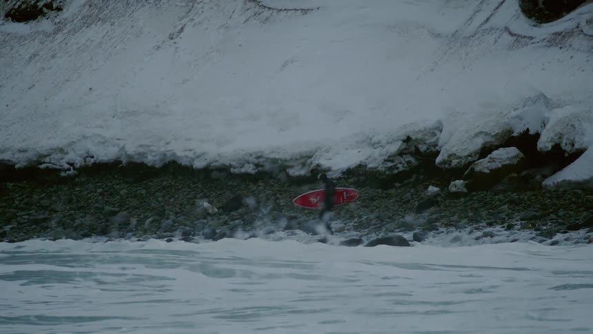 A person carrying a red surfboard walks along a rocky snow covered shoreline with waves crashing in the foreground