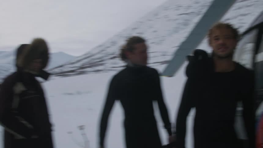 A group of men wearing black wetsuits unloading surfboards from a helicopter on a snowy landscape