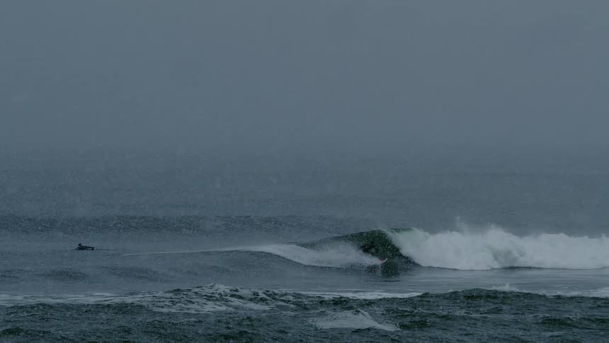 Surfer wearing a wetsuit rides a wave into shore as snow falls