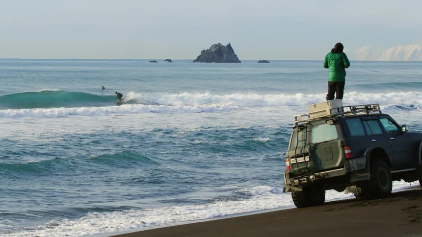 A photographer stands on top of a green suv and takes photos of surfers from an ocean coast