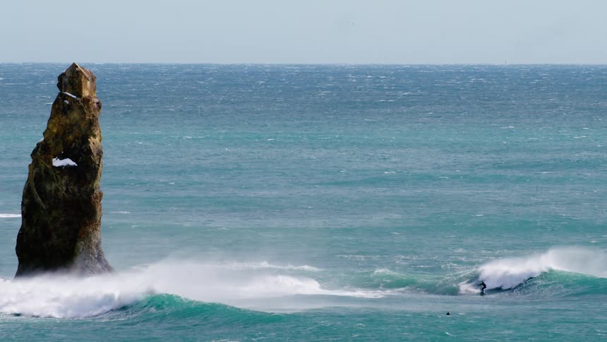 A surfer rides a wave near a tall rocky sea stack in the ocean