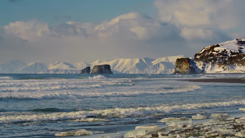 Waves roll in on an icy coast with snow covered mountains in the distance