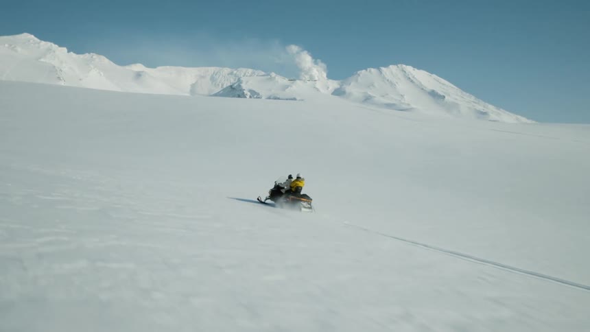 A lone snowmobile traverses a vast snowcovered landscape with a steaming volcano erupting in the distance