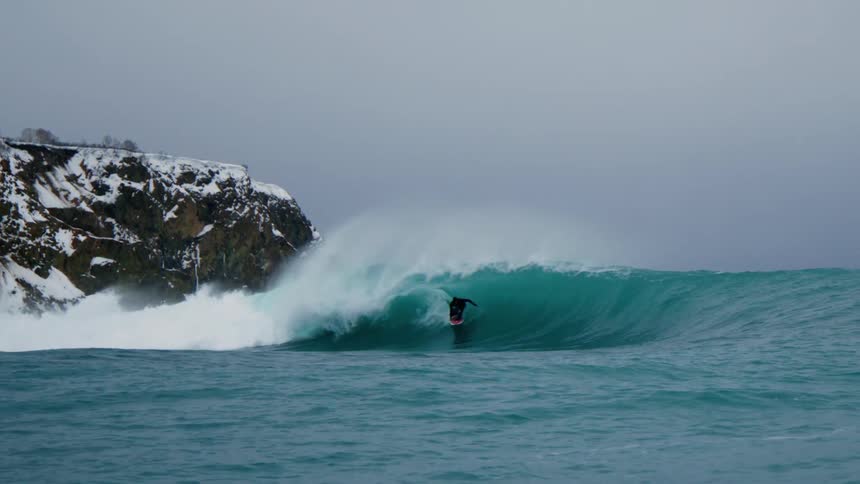 Surfer rides through the barrel of a wave cresting and crashing near a snowy promontory on an overcast winter day
