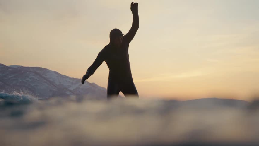 A man wearing a wetsuit rides a wave with snowy mountains in the background as the sun sets