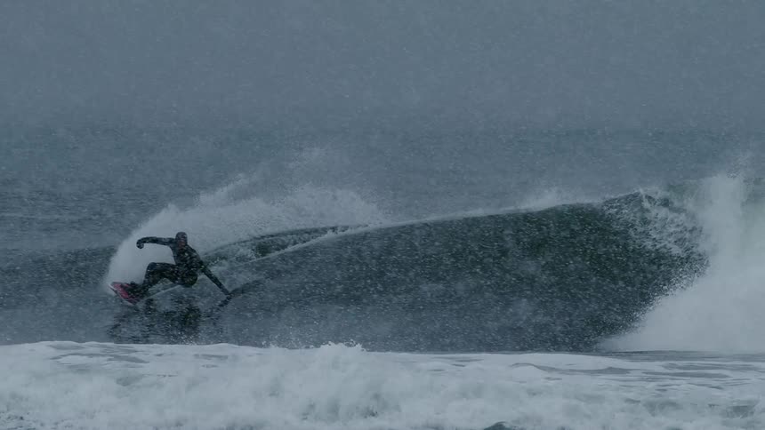 Water splashes over a surfer wearing a wetsuit as he rides a big wave into shore and snow falls