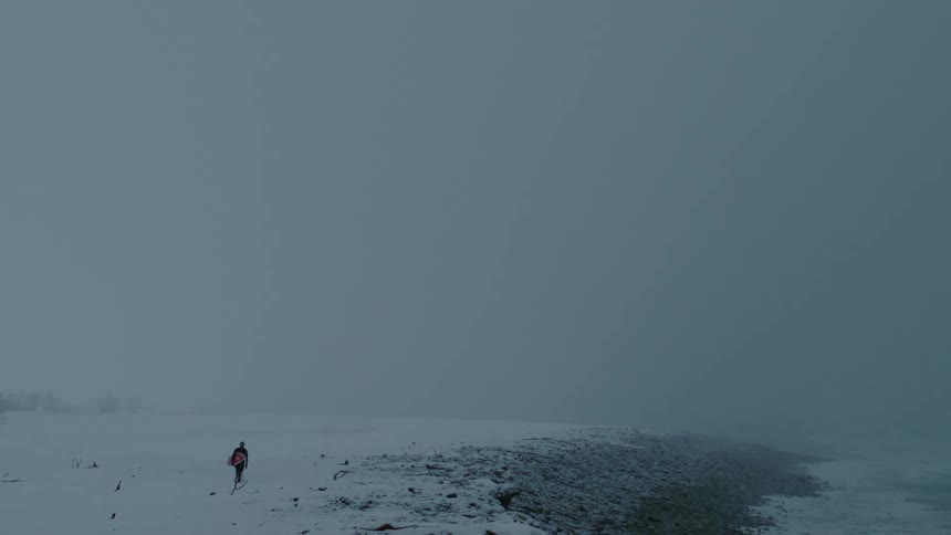 Person walking with a surfboard in a seaside setting on a snowy day
