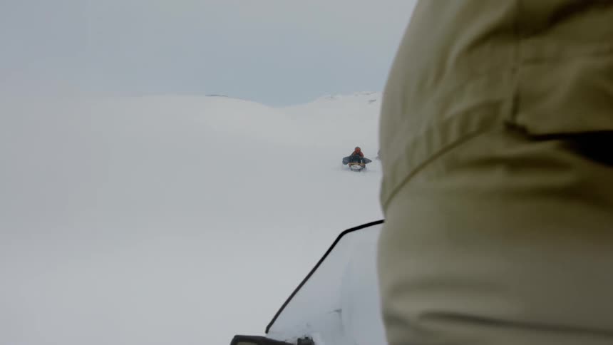 A person riding a snowmobile navigates through a snowy mountainous landscape with another snowmobile and rider visible in the distance