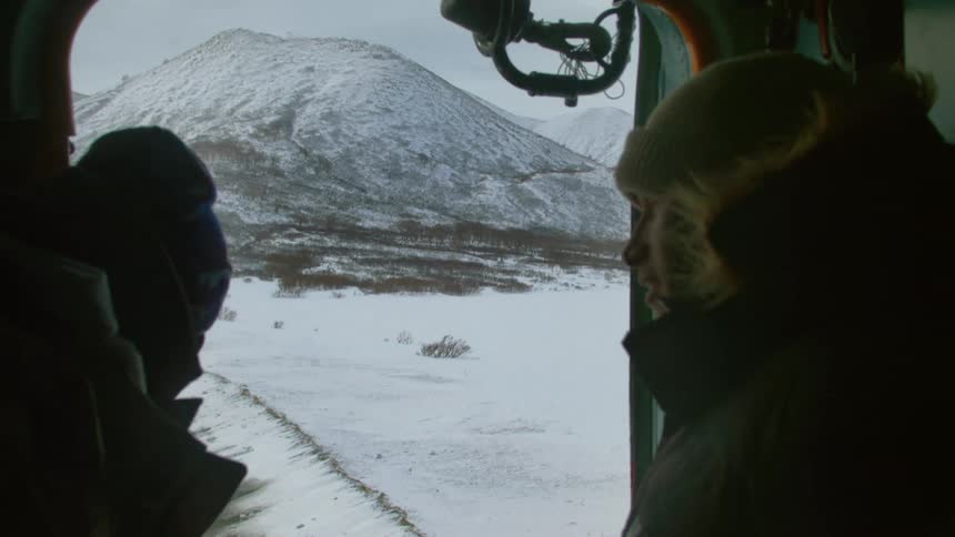 Two people look out from an open doorway of a helicopter at a snowy landscape with mountains in the background