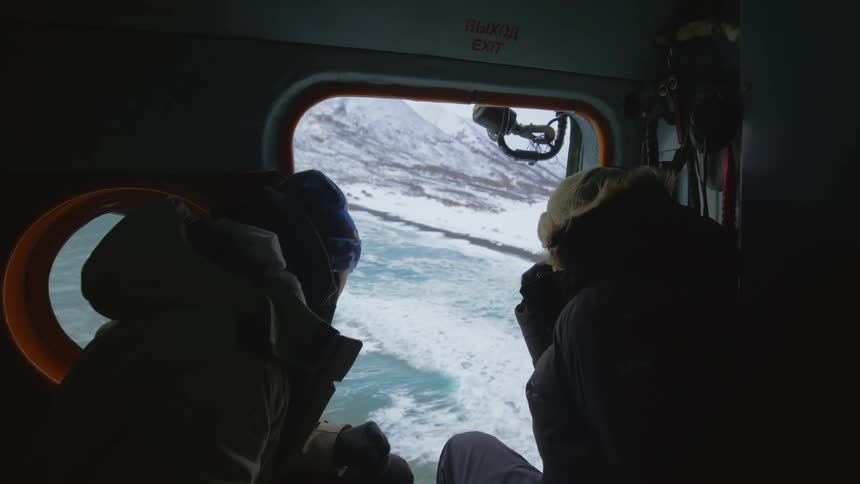 Two people inside a helicopter looking at the door flying above the sea with snow