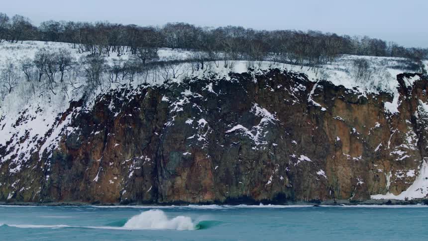 A wave crashes against a rocky shoreline near a snowdusted cliff face