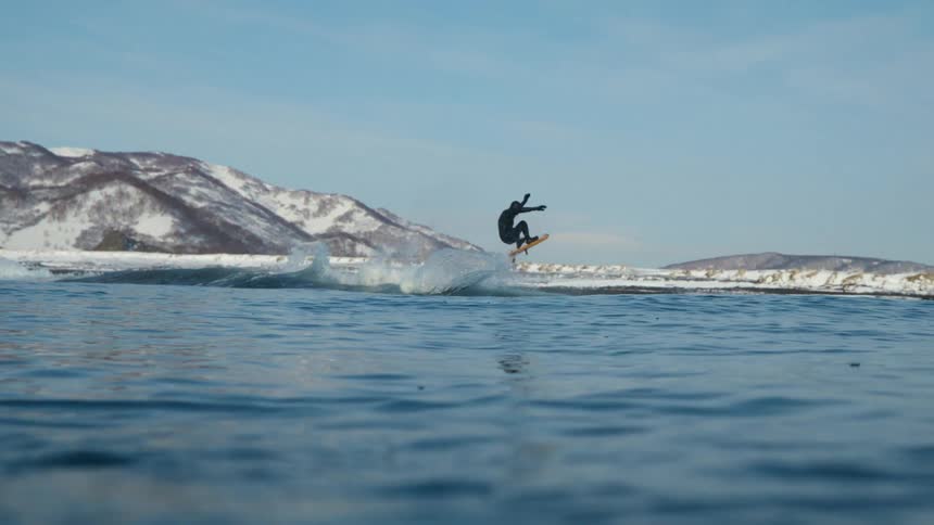 Surfer wearing a wetsuit leaps into the air on his surfboard with snow covered mountains along the coast