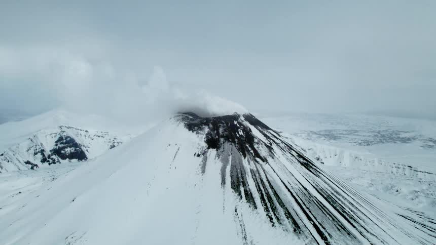 A plume of smoke rises from the snow capped peak of a volcano creating a dramatic scene against the cloudy sky