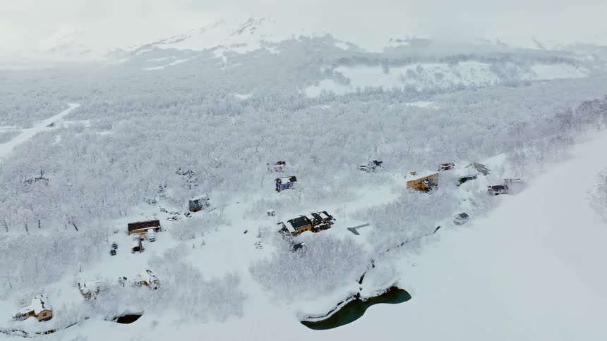 A picturesque winter landscape featuring a small village nestled amidst a blanket of snow with a frozen lake and snowcapped mountains in the background
