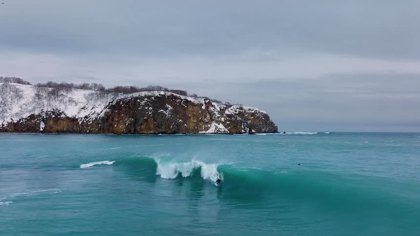 A surfer rides a wave near the coast close to a rugged snow capped cliff