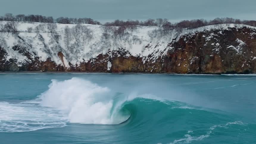 Several surfers are riding large waves in the ocean with a snowy cliff overlooking the scene