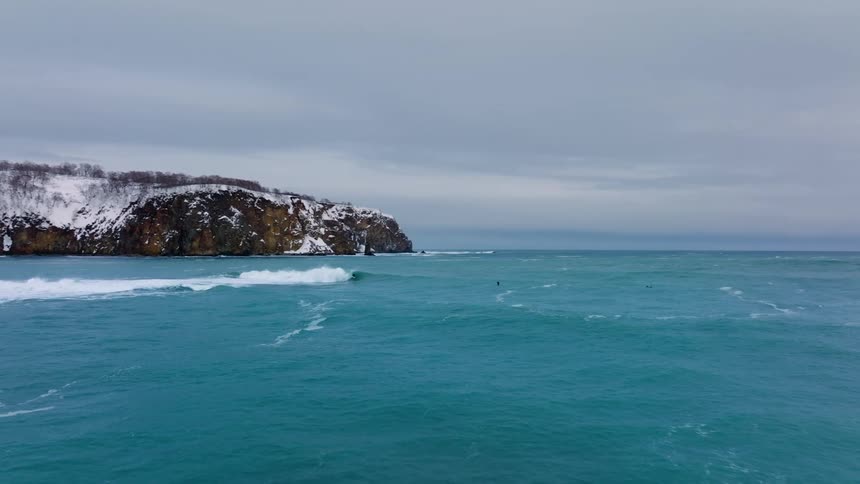 A surfer rides a wave in the ocean near a rocky cliff with snowcovered trees